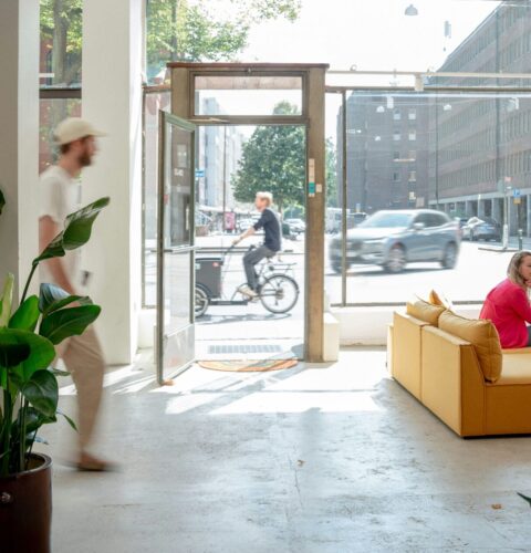 Bright office interior with large windows, a mustard-yellow couch, plants, a person walking, and others working or socializing.