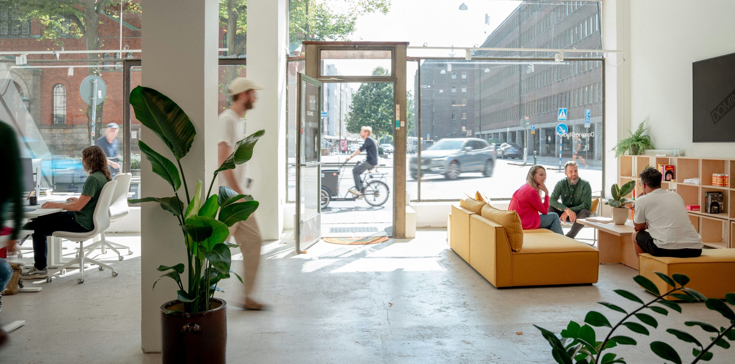Bright office interior with large windows, a mustard-yellow couch, plants, a person walking, and others working or socializing.
