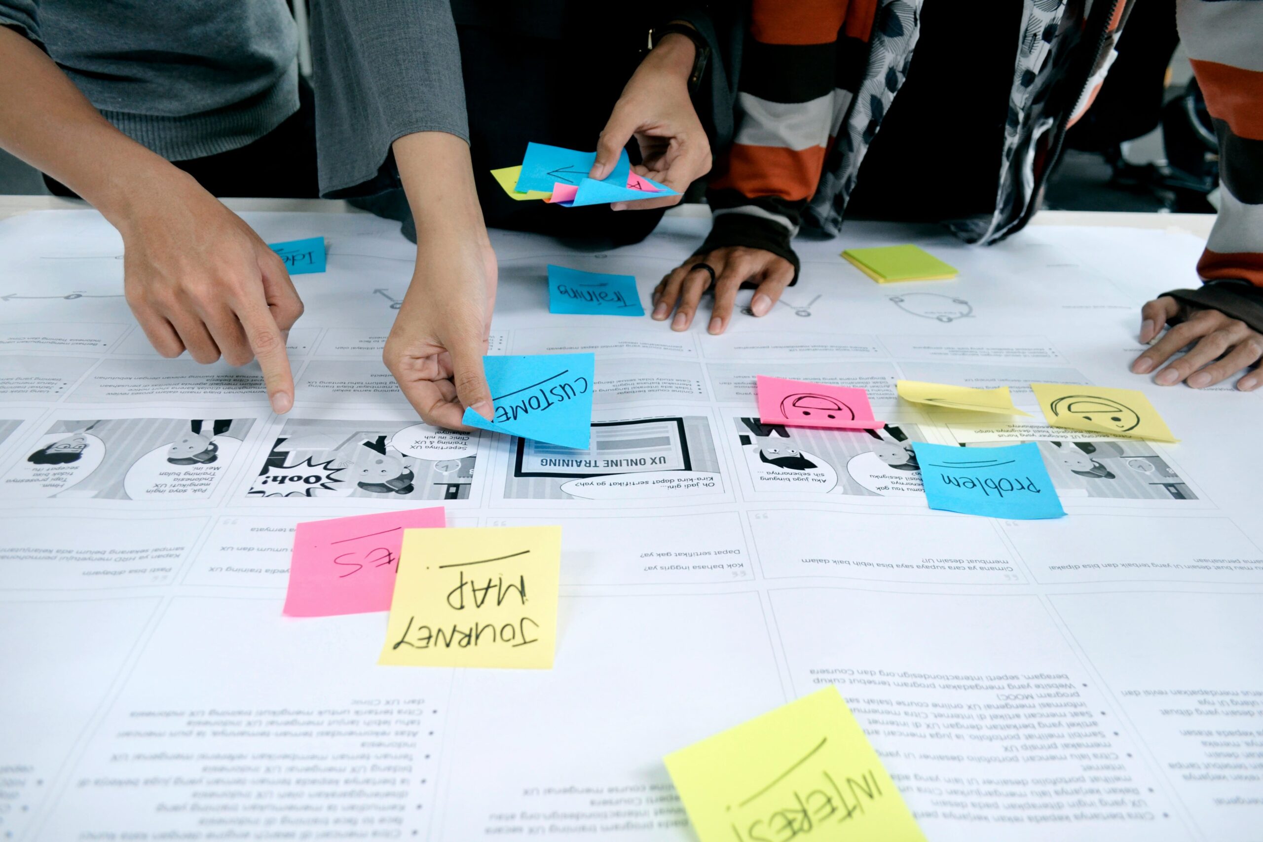 Hands of four people arranging colorful sticky notes labeled with words like "CUSTOMER," "JOURNEY MAP," and "PROBLEM" on a large paper diagram.